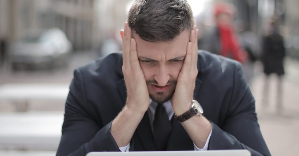 A man in a suit looks stressed while working on a laptop outdoors.