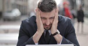 A man in a suit looks stressed while working on a laptop outdoors.