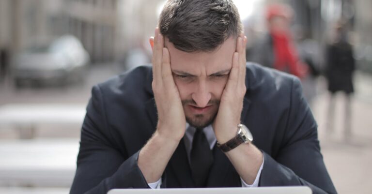 A man in a suit looks stressed while working on a laptop outdoors.