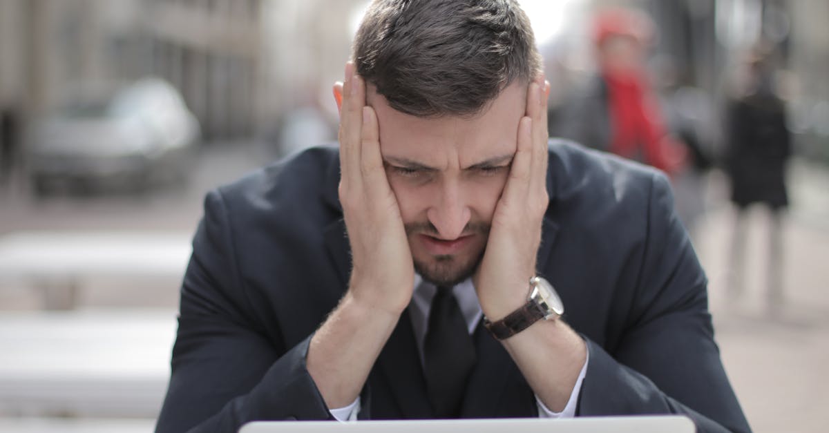 A man in a suit looks stressed while working on a laptop outdoors.